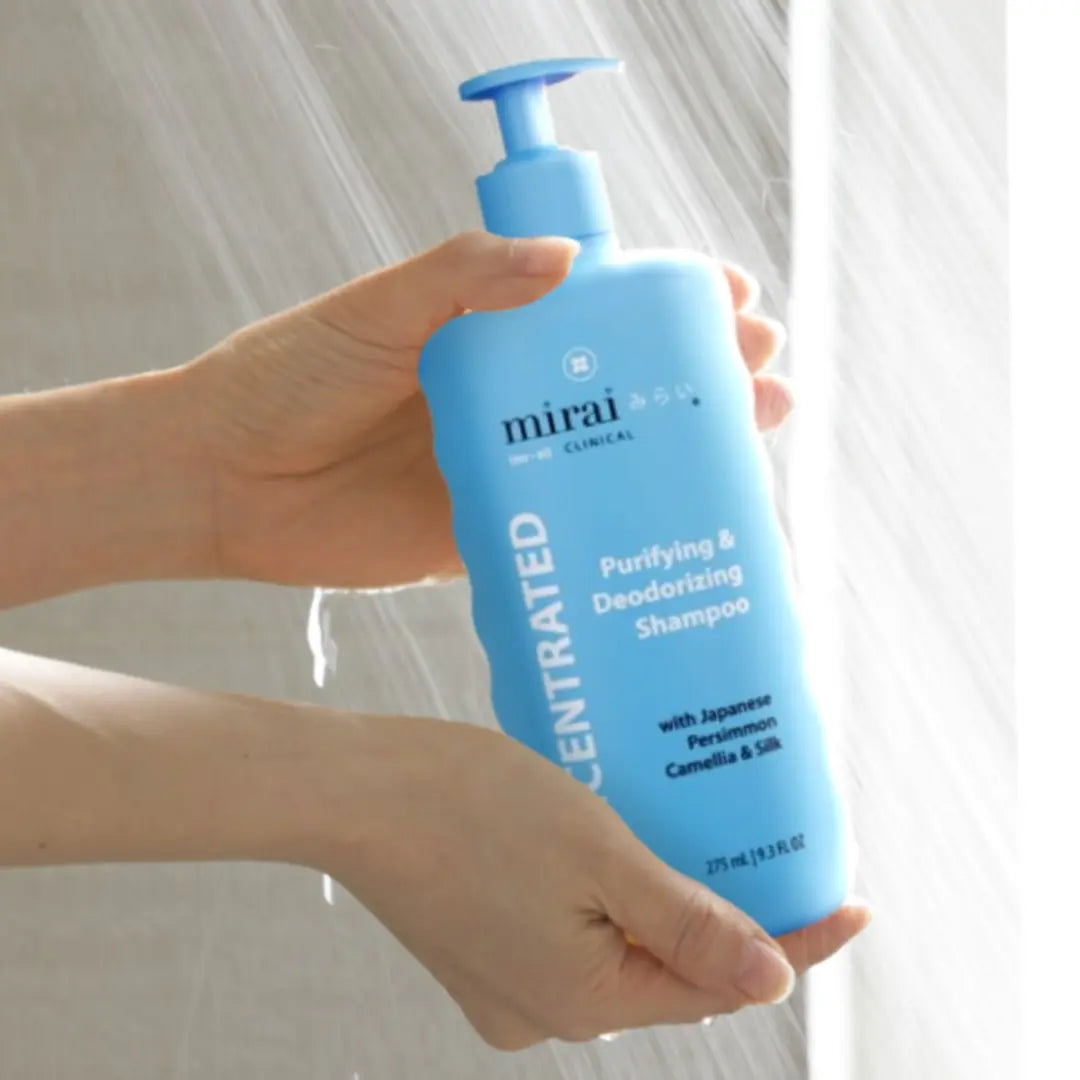 Hand holding Mirai Clinical Purifying & Deodorizing Shampoo under a shower, featuring Japanese persimmon and camellia to cleanse and nourish hair.