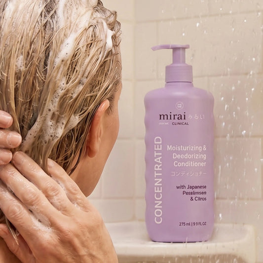 Person washing hair in the shower beside Mirai Clinical deodorizing conditioner with Japanese persimmon, designed to help reduce aging odor and moisturize hair.