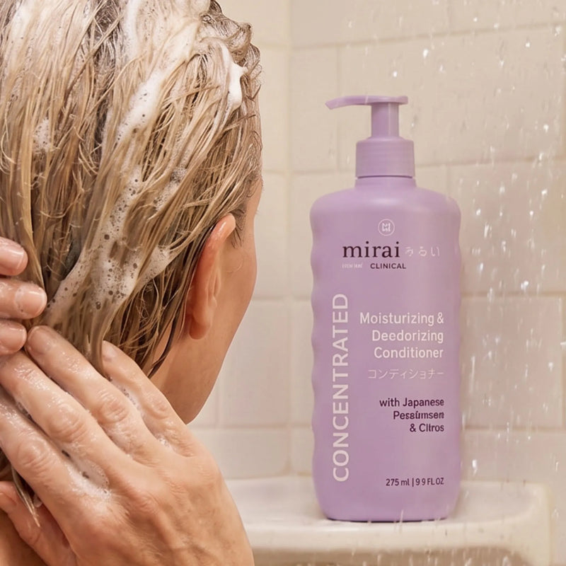 Person washing hair in the shower beside Mirai Clinical deodorizing conditioner with Japanese persimmon, designed to help reduce aging odor and moisturize hair.
