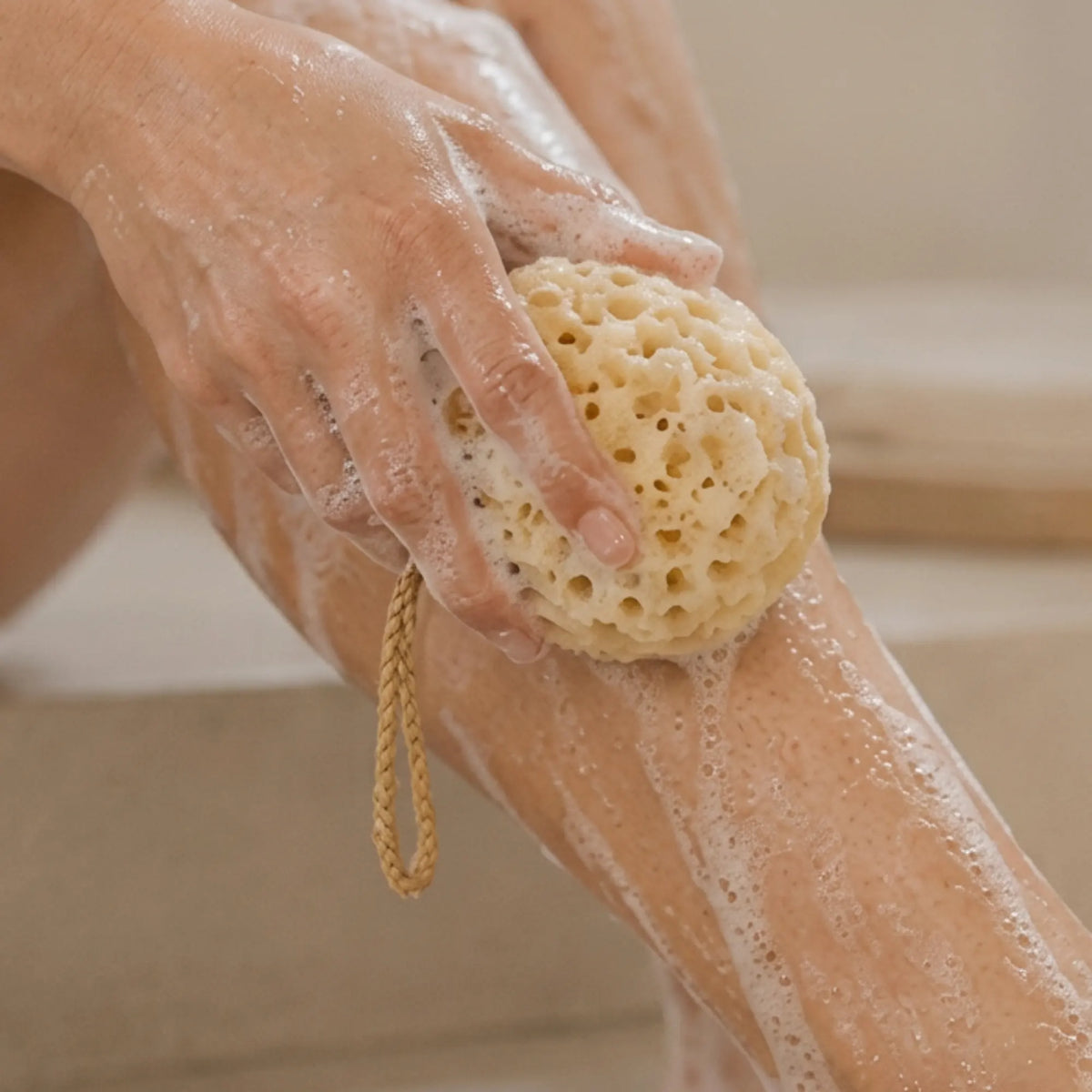 A close-up of a person washing their leg with a natural beige loofah sponge that has a braided loop handle, covered in white soap foam.