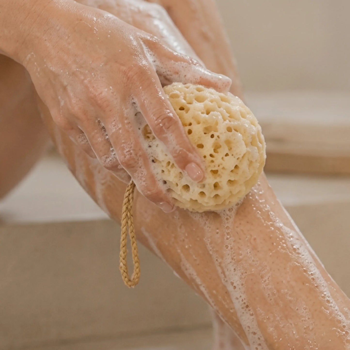A close-up of a person washing their leg with a natural beige loofah sponge that has a braided loop handle, covered in white soap foam.