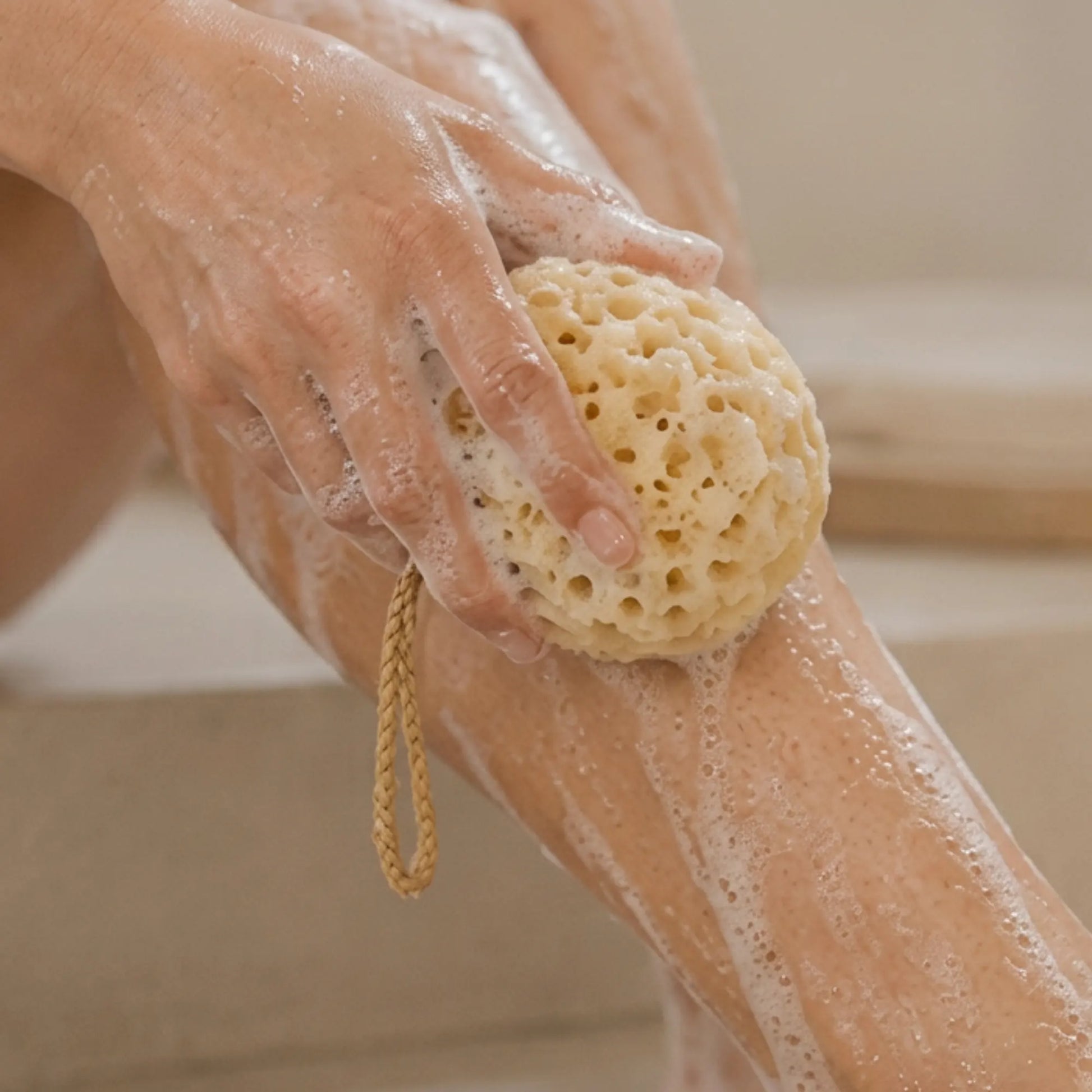 A close-up of a person washing their leg with a natural beige loofah sponge that has a braided loop handle, covered in white soap foam.