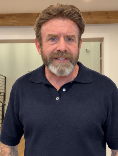 A man with a graying beard and brown hair wearing a dark navy blue polo shirt, standing indoors.

