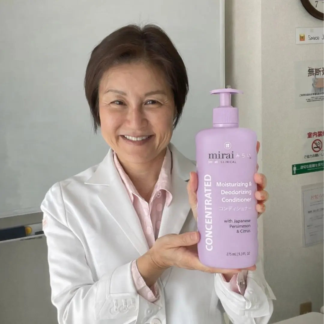 Smiling woman in a white lab coat holding a large bottle of Mirai Clinical Moisturizing & Deodorizing Conditioner with Japanese Persimmon and Citrus.