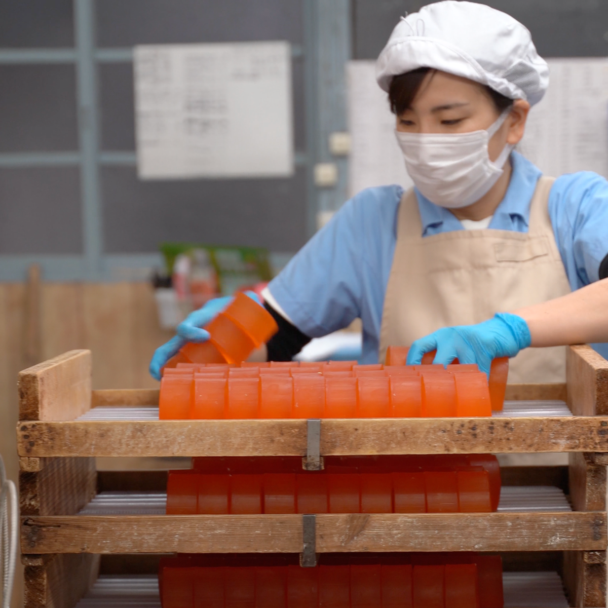 Worker in mask and gloves arranging orange persimmon soap bars on wooden drying racks in a Japanese soap factory