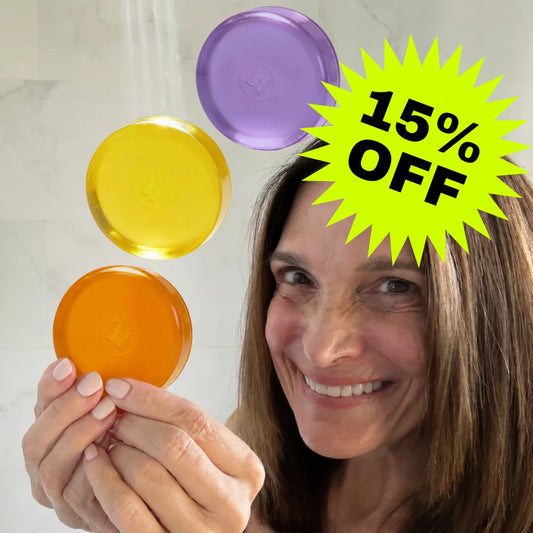 Woman holding 3 Mirai Clinical Nonenal soap bars — Lavender (purple), Citrus (yellow), and Regular (orange) — with 15% off promotional badge