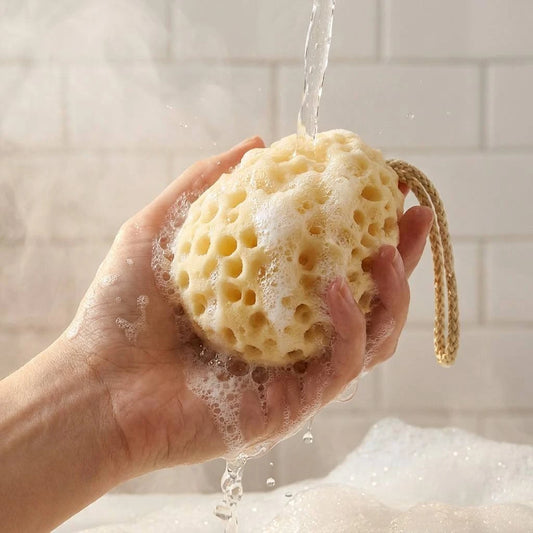 A hand holding a porous natural sponge bath scrubber under running water, with soap foam and bubbles visible against a light tiled bathroom background.