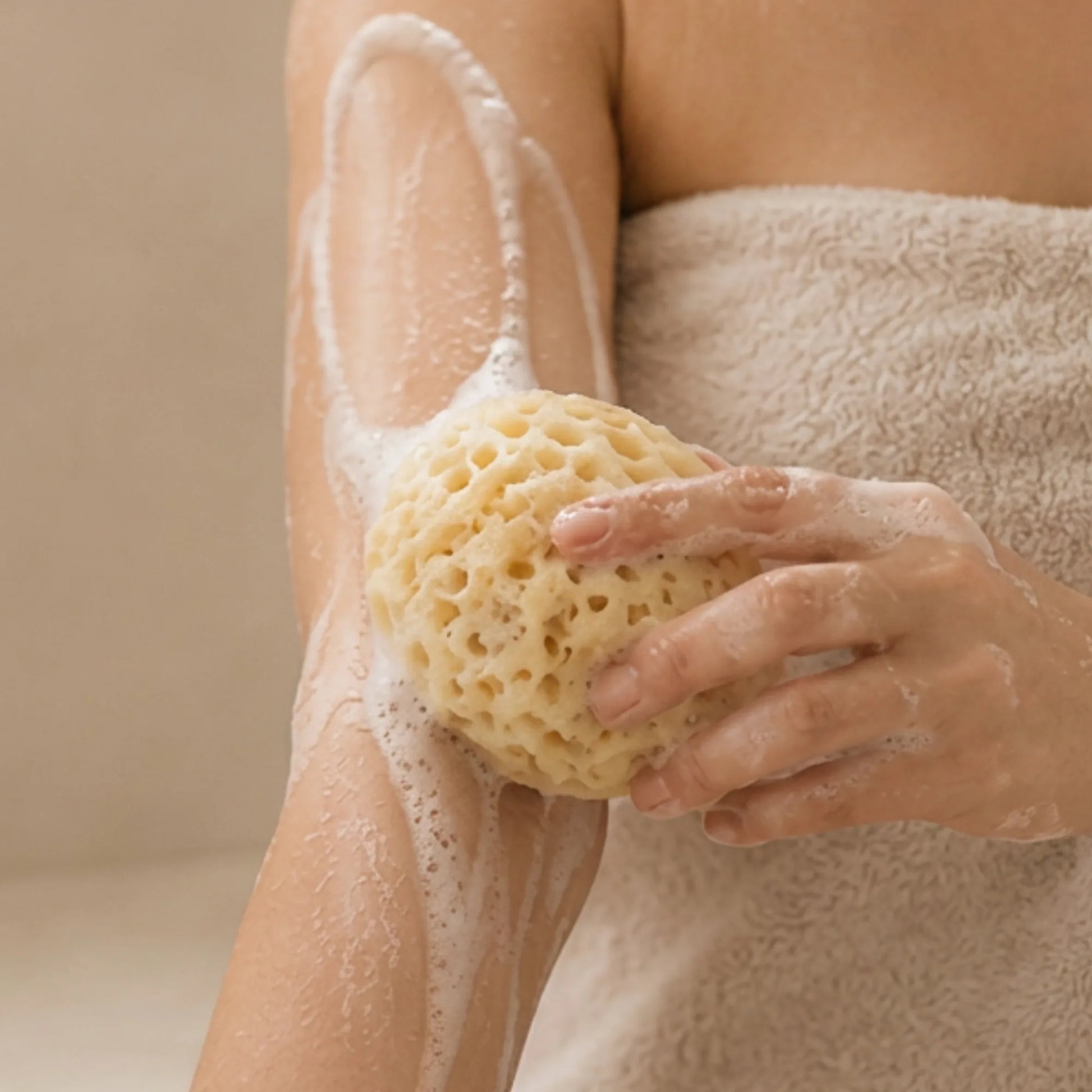 A person wrapped in a beige towel washes their arm with a natural beige sea sponge, with white soap lather visible on the skin.