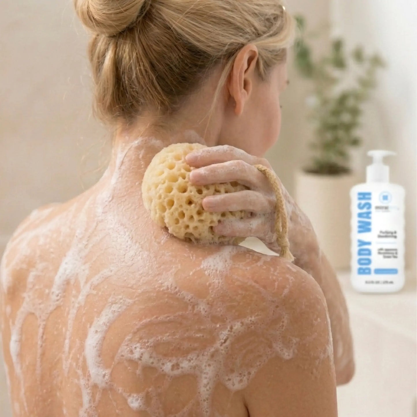 A woman with sapy skin is shown from behind, using a natural sponge on her shoulder in a bathroom setting. A bottle labeled 'BODY WASH' is visible in the background.